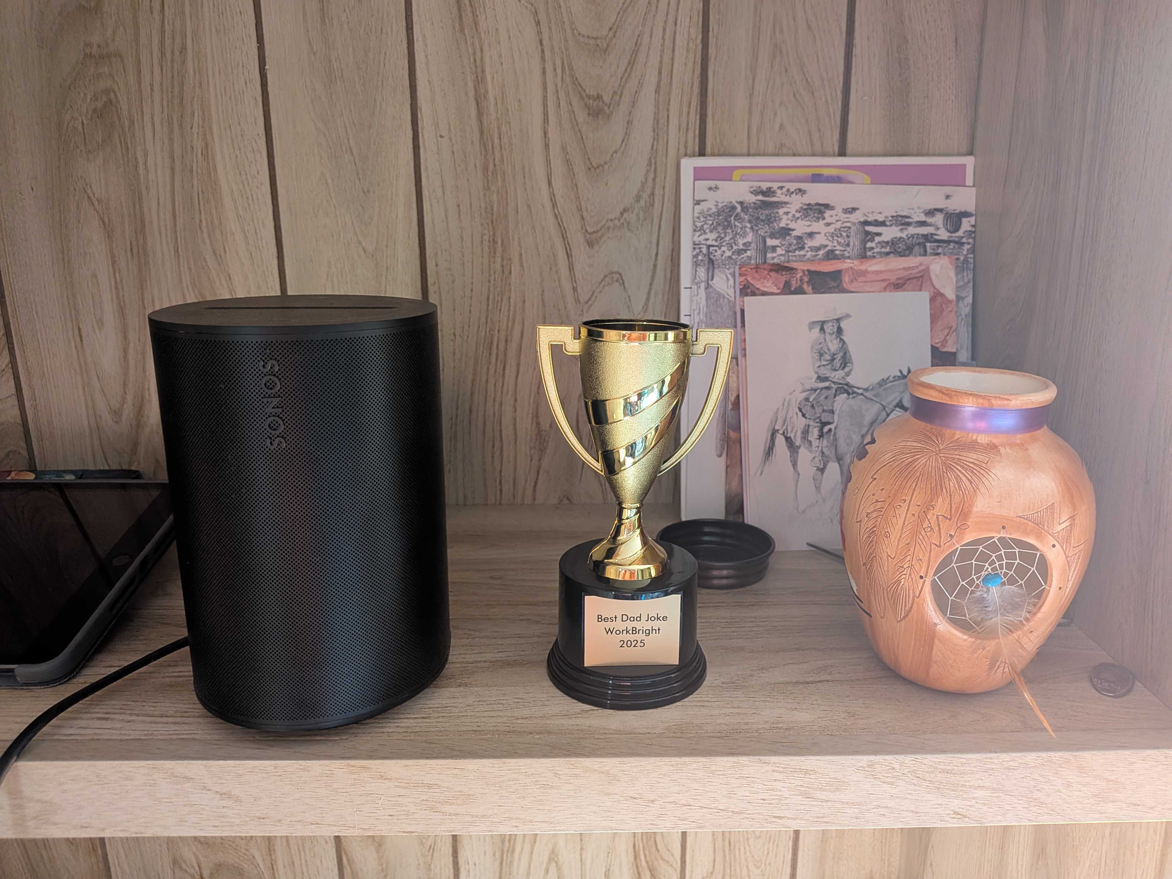 Gold trophy engraved "Best Dad Joke WorkBright 2025" on a wooden shelf next to a black Sonos speaker and a painted clay vase with a dreamcatcher. Gold trophy engraved "Best Dad Joke WorkBright 2025" on a wooden shelf next to a black Sonos speaker and a painted clay vase with a dreamcatcher.