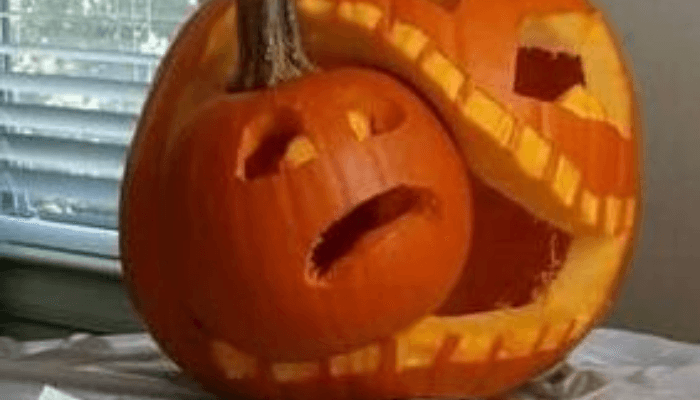 A carved pumpkin with a wide mouth appears to be eating a smaller whole pumpkin, both lit from within on a paper-covered table. A carved pumpkin with a wide mouth appears to be eating a smaller whole pumpkin, both lit from within on a paper-covered table.