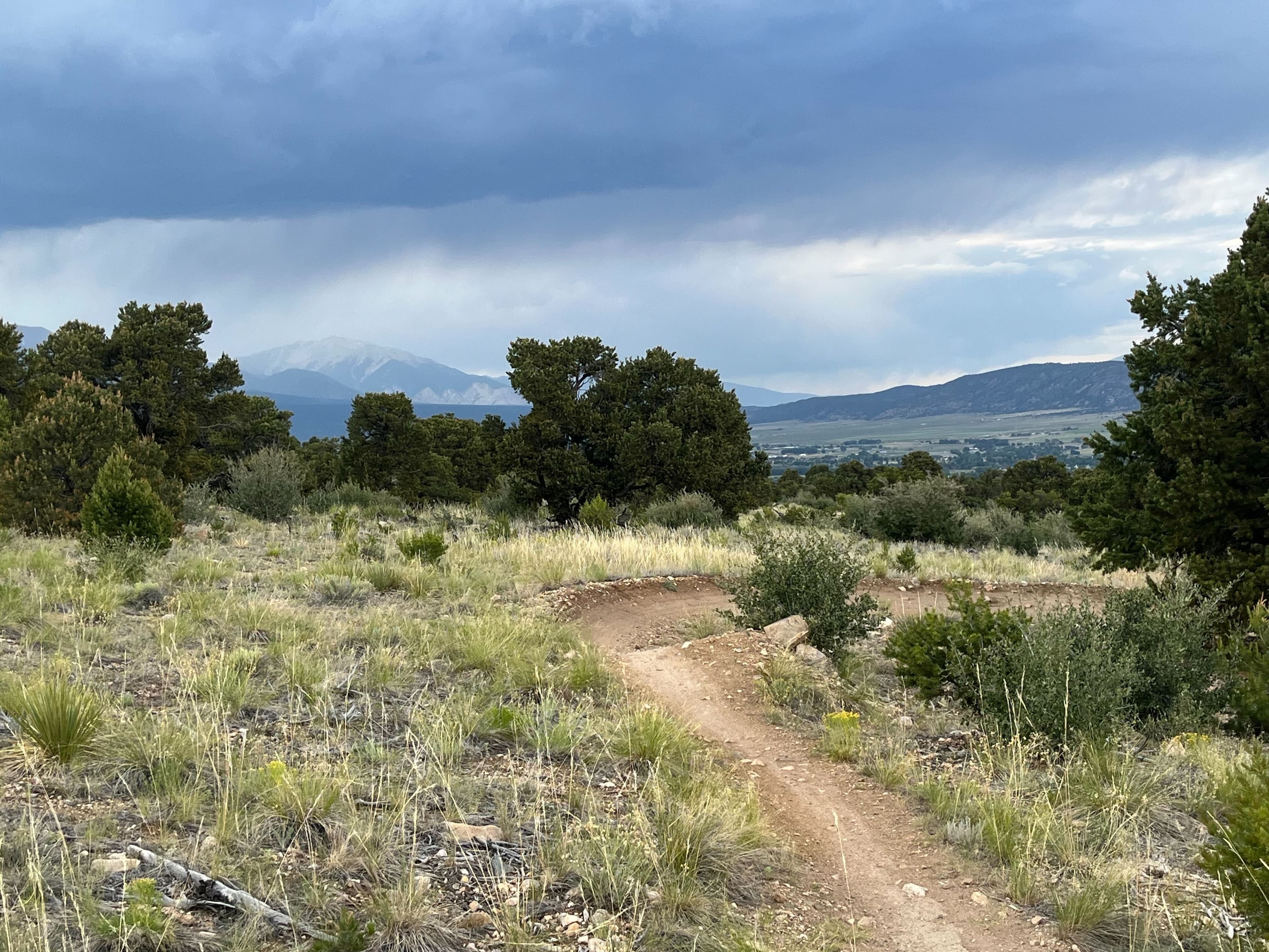 A narrow dirt singletrack trail winds through scrubland and juniper toward a snow-dusted mountain range under heavy gray storm clouds. A narrow dirt singletrack trail winds through scrubland and juniper toward a snow-dusted mountain range under heavy gray storm clouds.