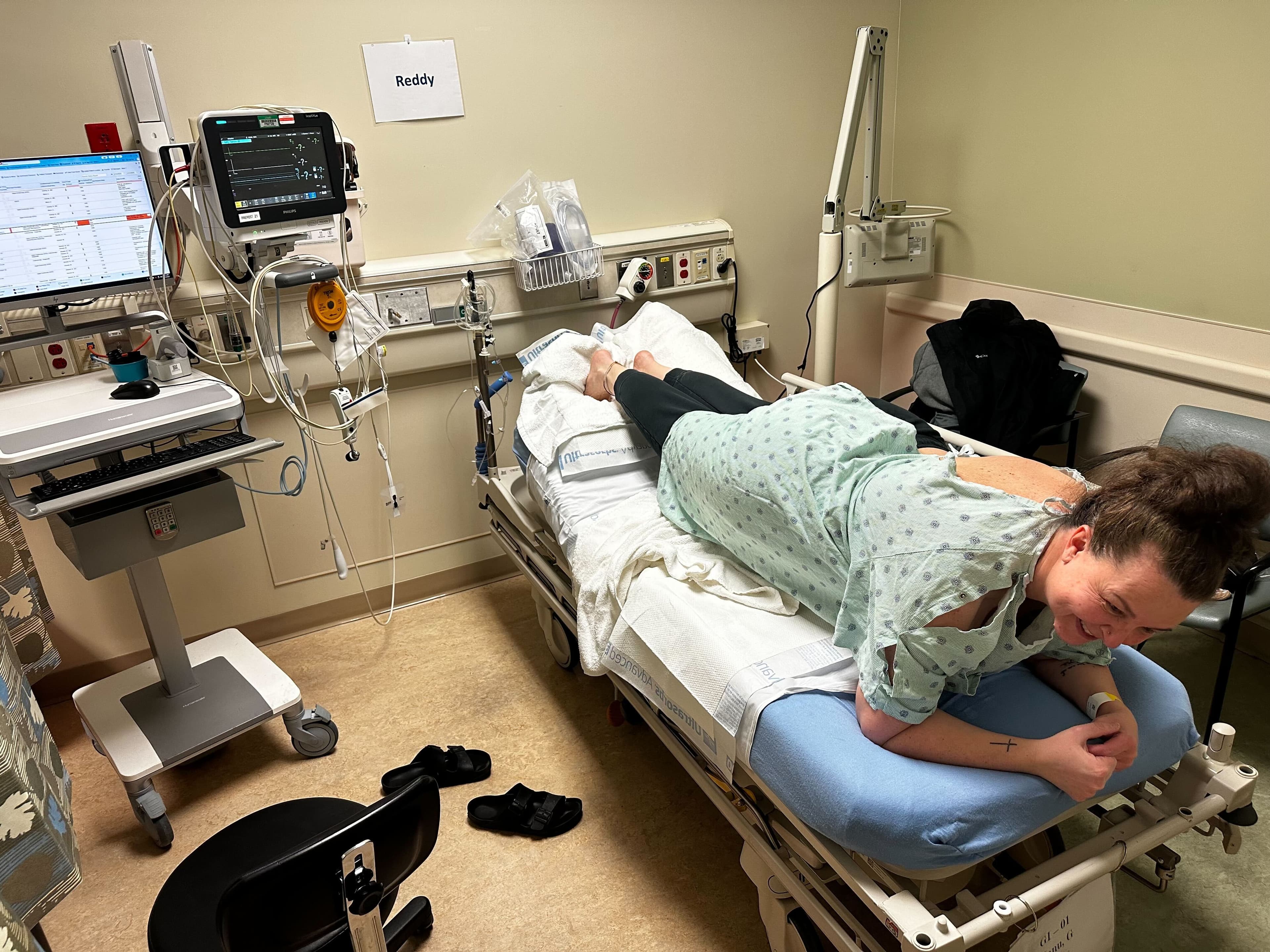 A person in a hospital-style gown braces in a plank position over a pillow on a bed, smiling through a coworker plank challenge. A person in a hospital-style gown braces in a plank position over a pillow on a bed, smiling through a coworker plank challenge.