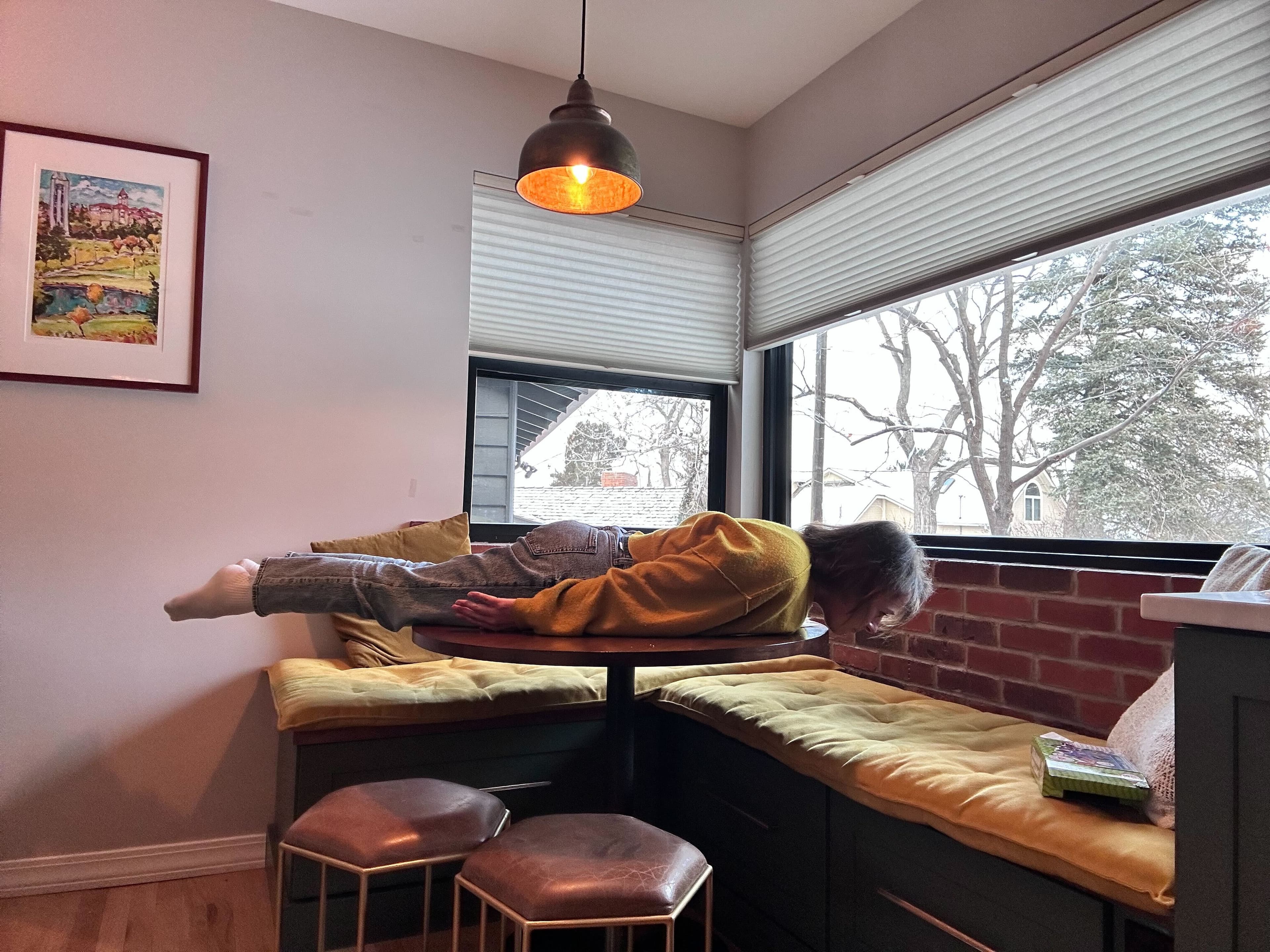 A person in a yellow sweater and jeans lies stiff and horizontal across a round breakfast-nook table beside windows showing a snowy backyard. A person in a yellow sweater and jeans lies stiff and horizontal across a round breakfast-nook table beside windows showing a snowy backyard.