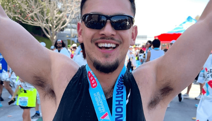 A man in a black tank top and sunglasses lifts his arms triumphantly at the LA Marathon finish, wearing the blue-ribboned "40 Years Running" medal. A man in a black tank top and sunglasses lifts his arms triumphantly at the LA Marathon finish, wearing the blue-ribboned "40 Years Running" medal.